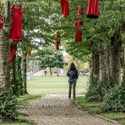 A young woman with a backpack walks a path underneath trees through the Red Dress Project installation in Spencer Smith Park